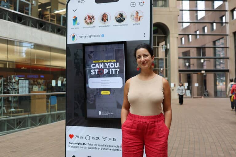 Commissioner Govender stands in the central Vancouver Public Library, wearing a sleeveless beige top and red pants. Behind her is a giant cellphone showcasing the Misinformation quiz.