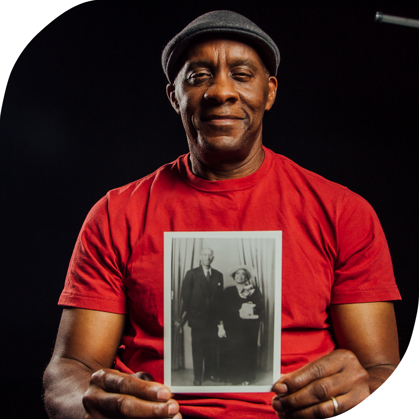 A photo of Anthon Brown, a Black man in his 60s who is wearing an orange shirt and hat. He is holding a black and white photo of his grandparents.