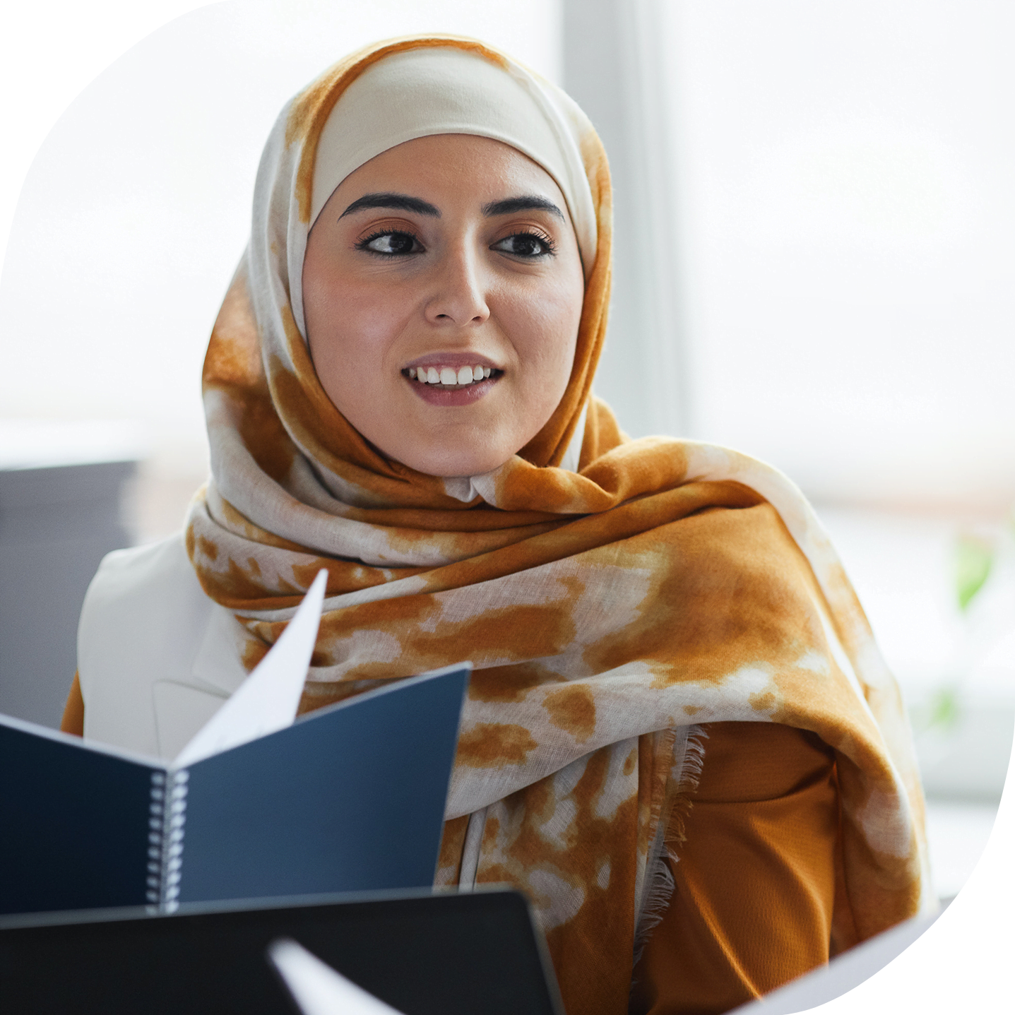 An Arab woman wearing a yellow and white hijab smiling at a colleague who is not pictured. She is flipping through a book and sitting in an office setting.