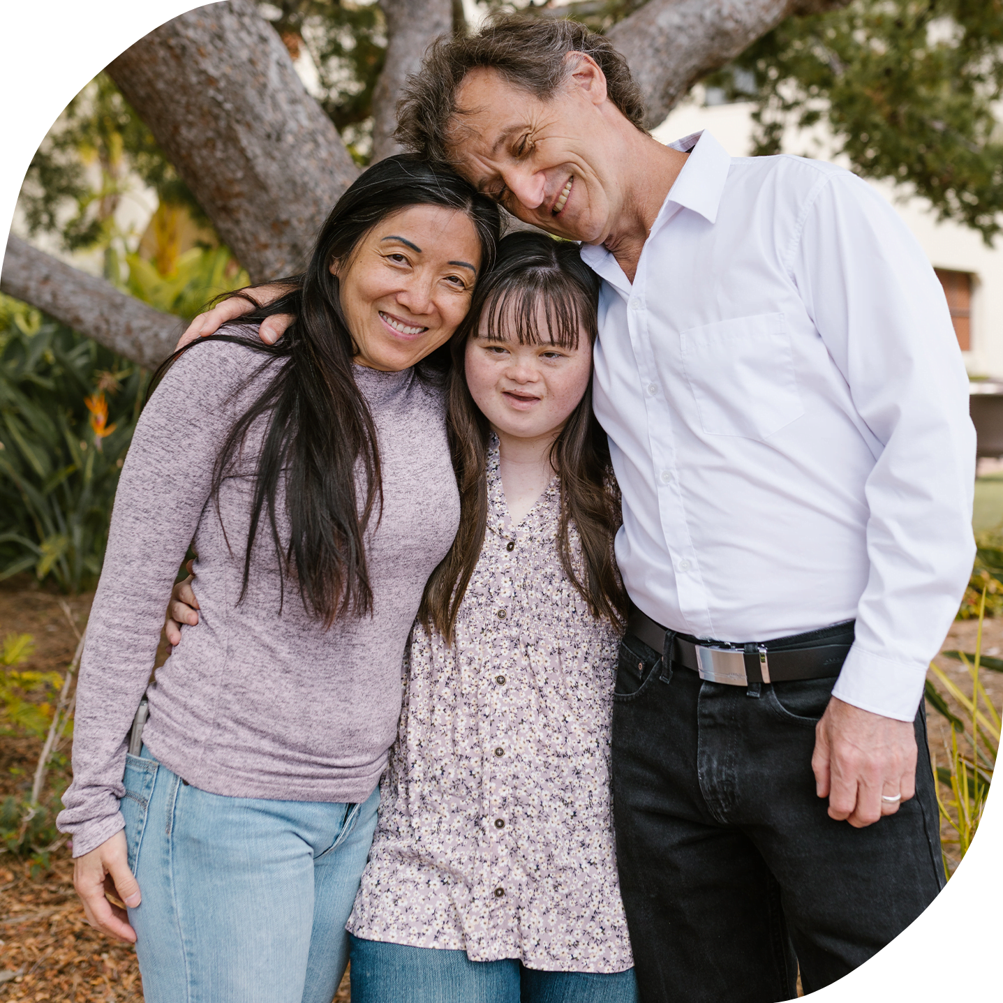 This image depicts a mixed race family with a mother and a father embracing their daughter who has Down's syndrome. All three are smiling and have expressions of joy.