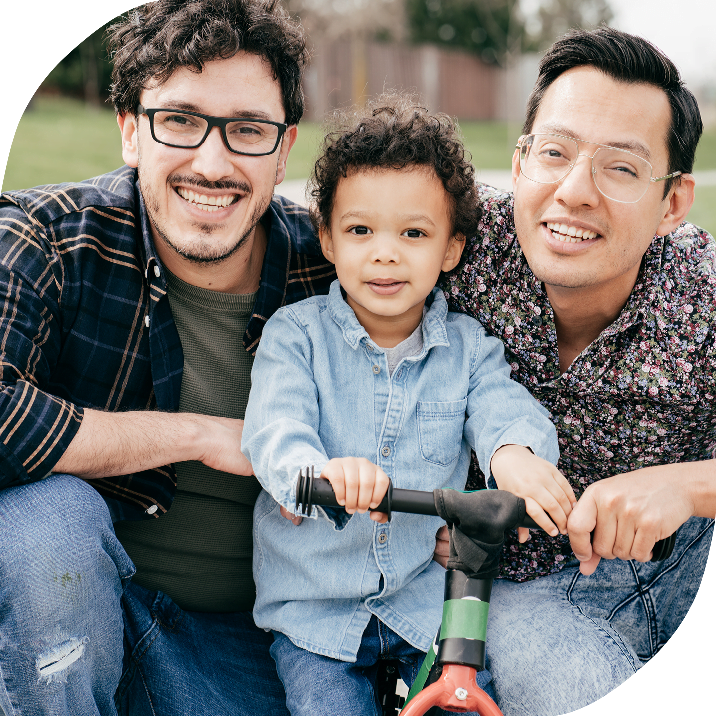 A young, mixed race LGBTQ2AS+ family where two dads are on either side of their young son who is holding the handlebars of a bicycle