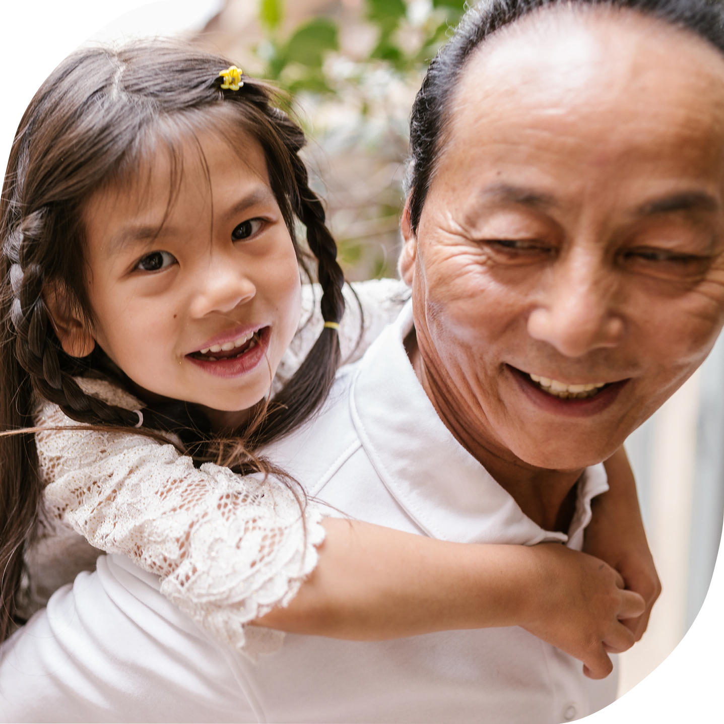 A smiling Asian father or grandfather is holding his young daughter on his back and looking lovingly in her direction. Her face is directed towards the camera and she is smiling. Her arms are wrapped around her relative's neck.