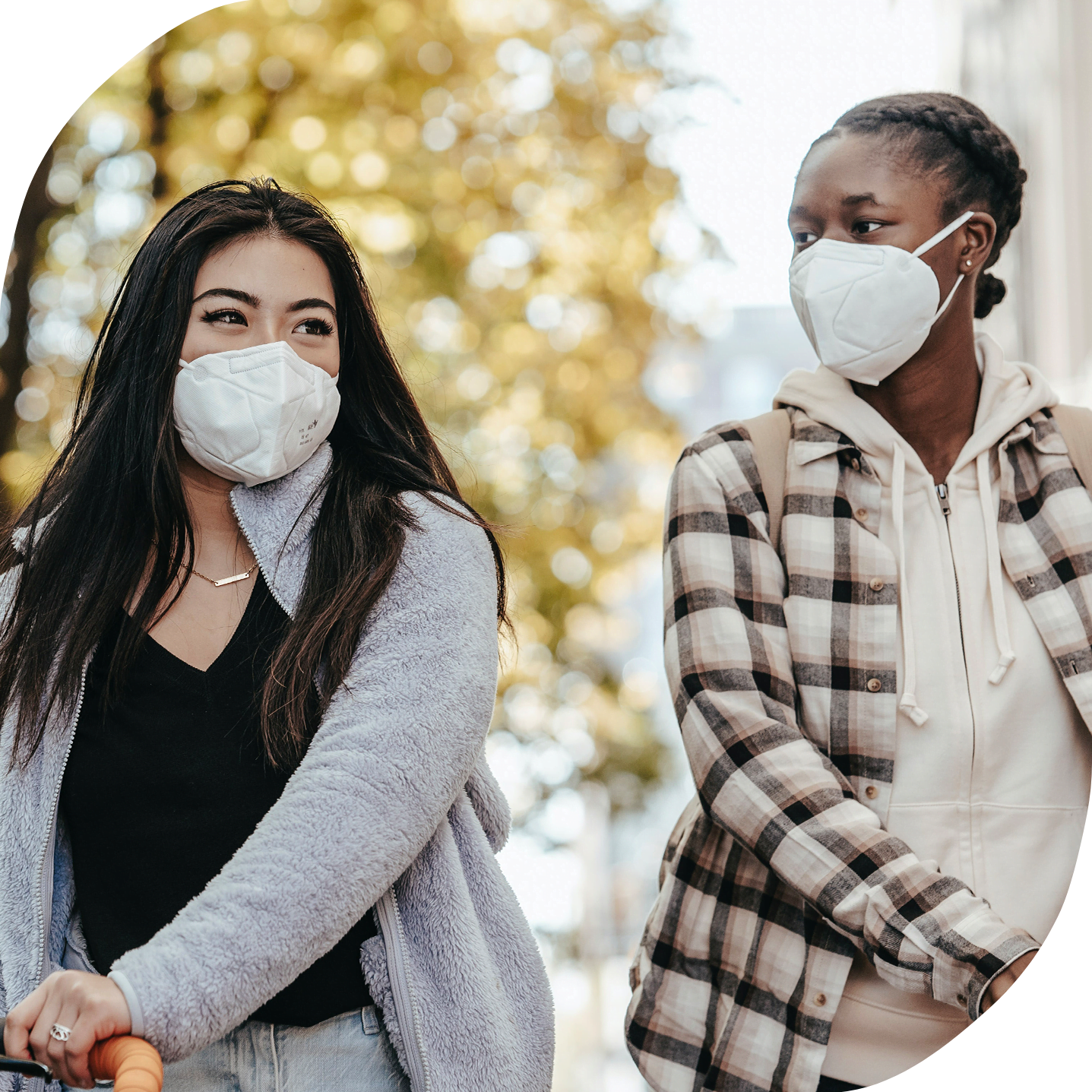Two diverse young women are walking their bikes along a sidewalk and wearing masks while in conversation with one another.