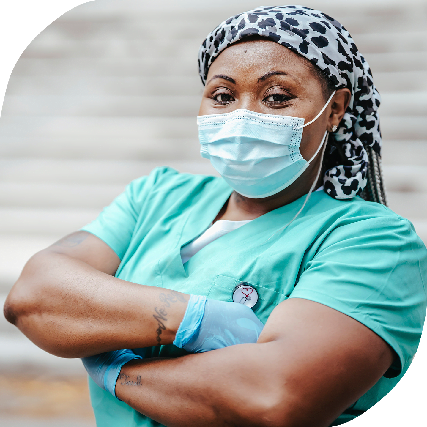 A Black woman wearing a mask and health care worker scrubs is looking into the camera.