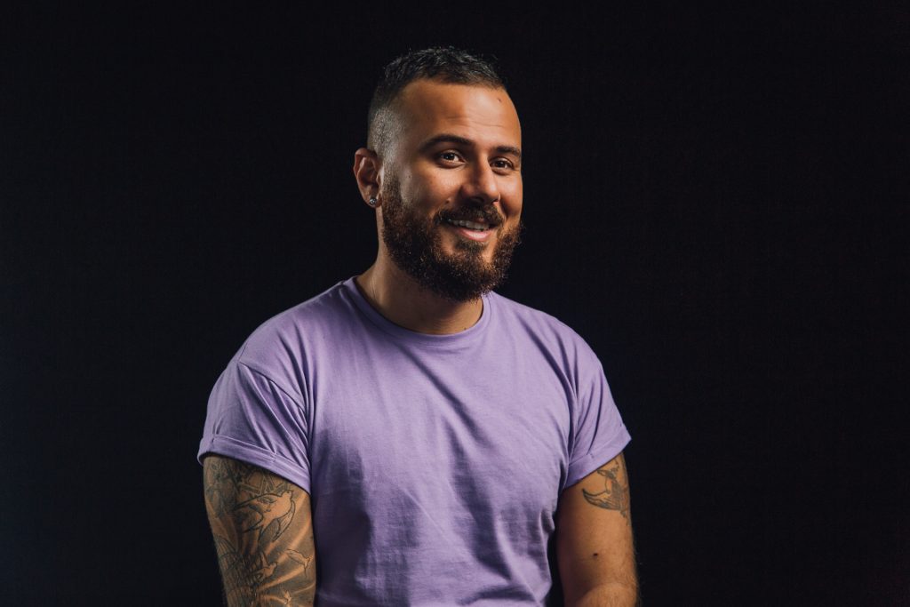 Danny Ramadan, a young Syrian man with short dark hair and a beard, is smiling at a studio. He has tattoos on his arms and is wearing a purple shirt.