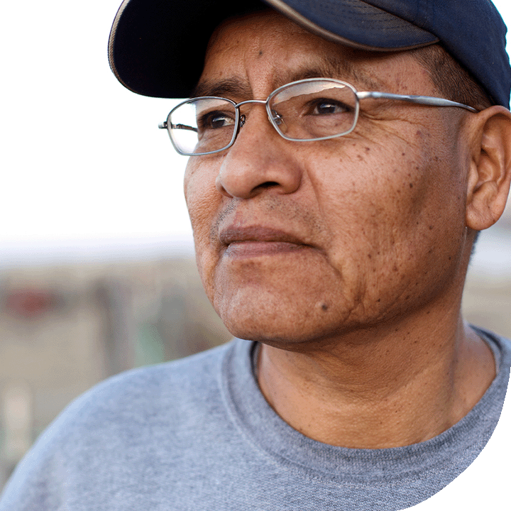 A middle-aged Indigenous man wearing a black cap, grey shirt and glasses looks into the distance.