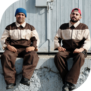 Two men wearing blue collar jumpsuits are sitting on a ledge smiling at the camera. The man on the left has medium skin tone, is more stout, and is wearing a blue toque. The man on the right has light-medium skin tone, dark facial hair, and is wearing a red cap.