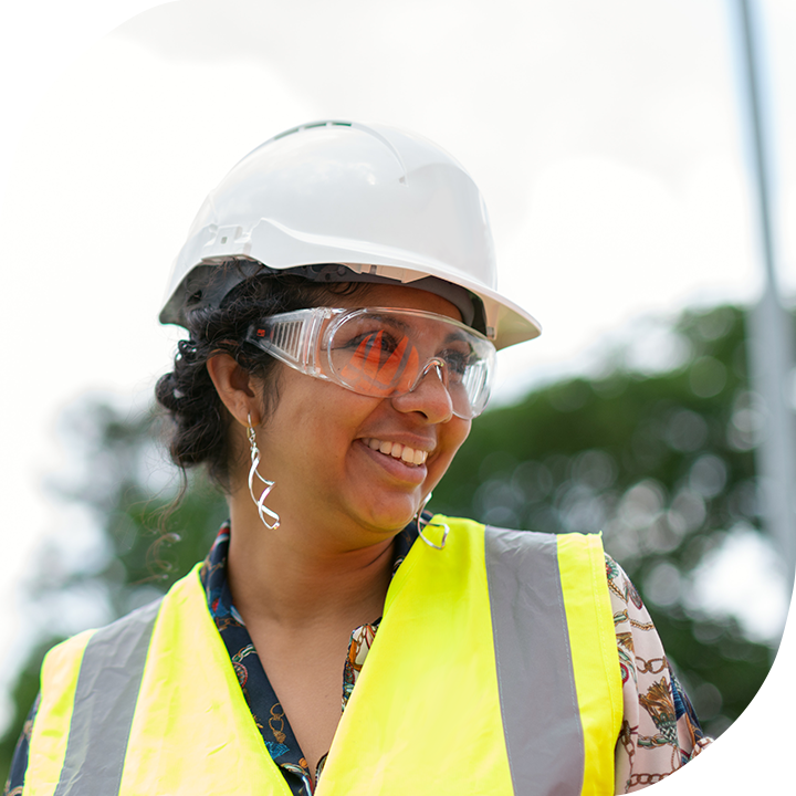 South Asian woman in a safety vest, earrings, and hard hat. She is smiling to the right.