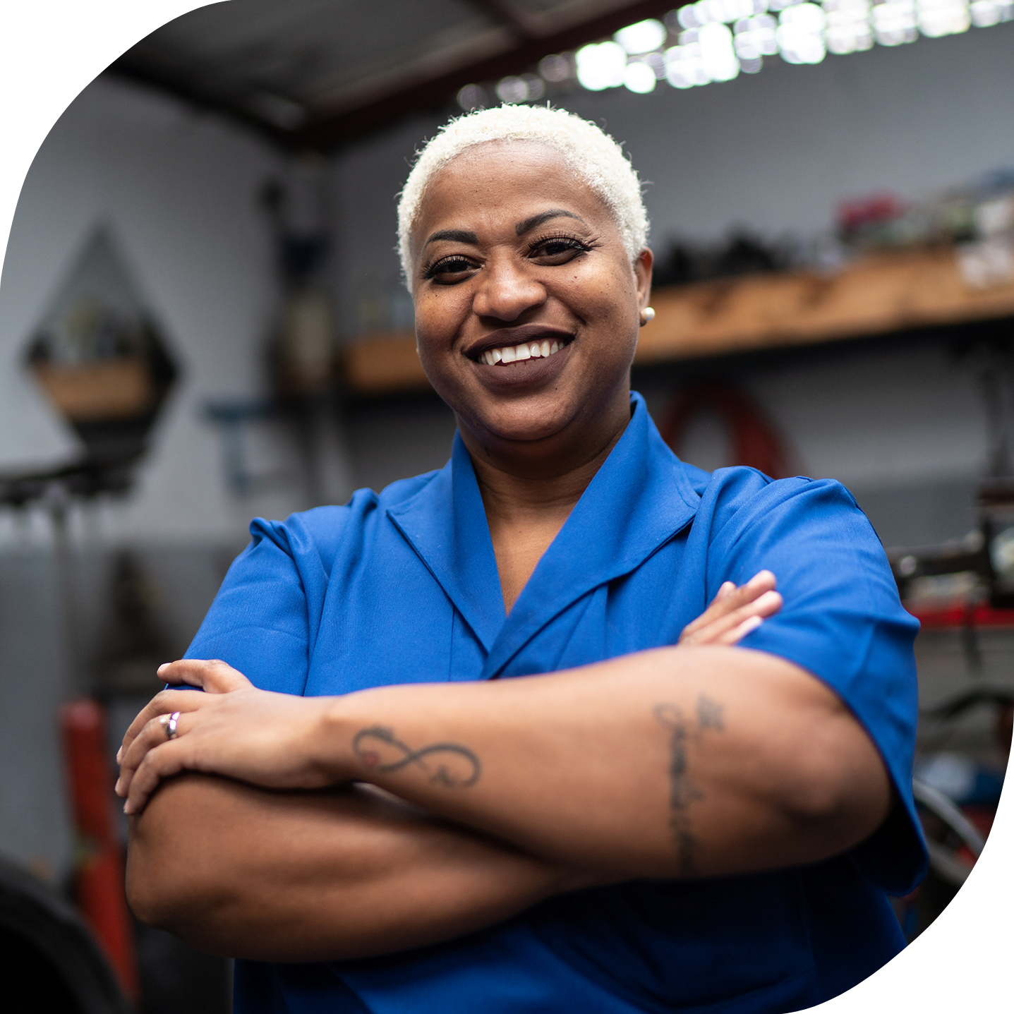 A Black woman with short blonde hair in a blue uniform has her arms crossed and is smiling. She is working at a auto car repair service center.