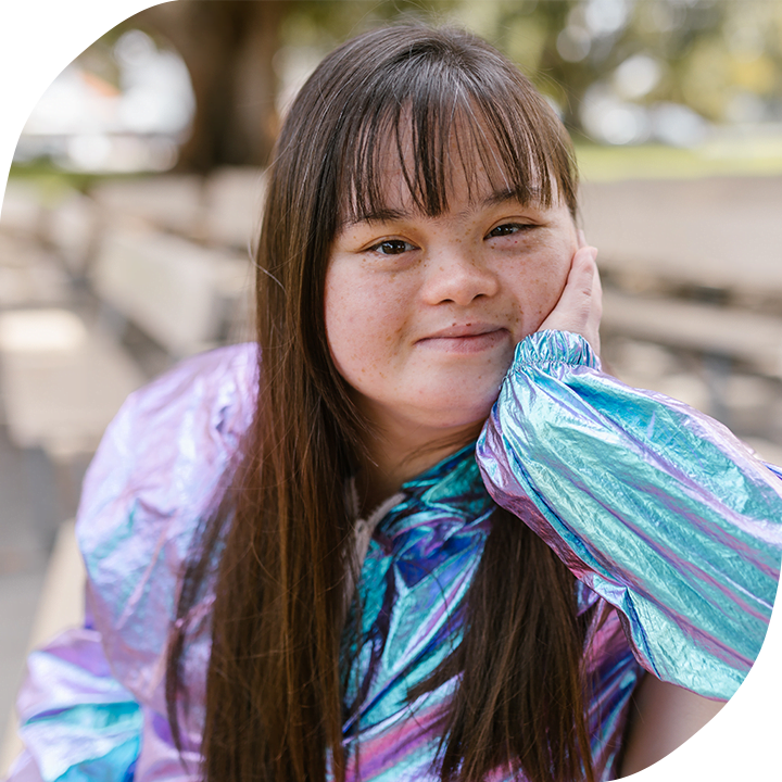 A light-skinned girl with down syndrome is sitting on a bench on a bright sunny day, smiling directly into the camera