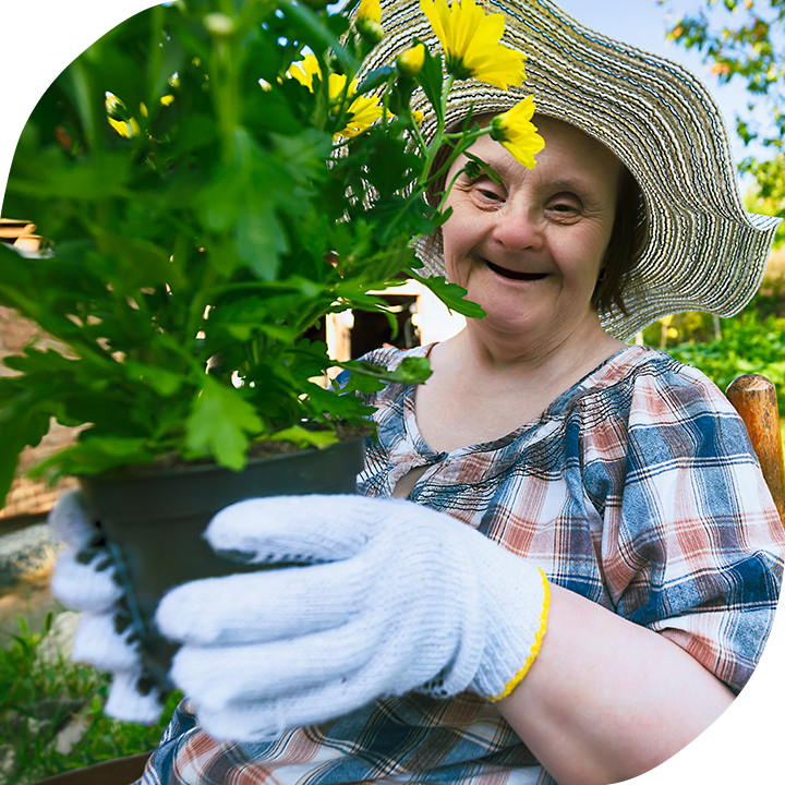 Older white woman with down syndrome wearing a sun hat and gardening gloves, showing off a pot of yellow flowers and smiling