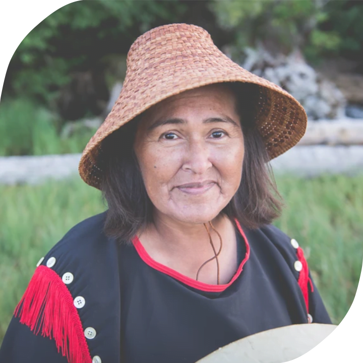 A woman from the Gitxaala Nation is wearing a black and red dress and a traditional woven hat. She has brown hair and is standing in a grassy field, smiling into the camera.