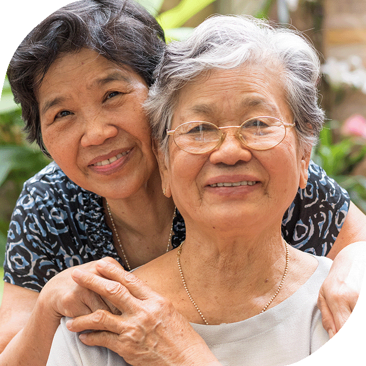 Two Asian grandmas hold each other, smiling. One has short black hair and the other has short grey wavy hair.