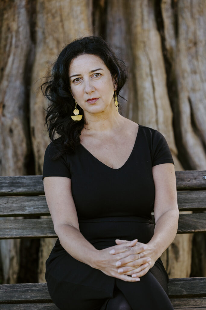 Commissioner Kasari Govender sits on a bench in front of a large red cedar tree. She has a light skin tone with black hair and wears a black v-neck shirt, black pants and gold earrings.