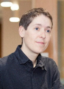 A photograph of Trish Garner who is white and has short brown hair. They are wearing a black button up dress shirt.