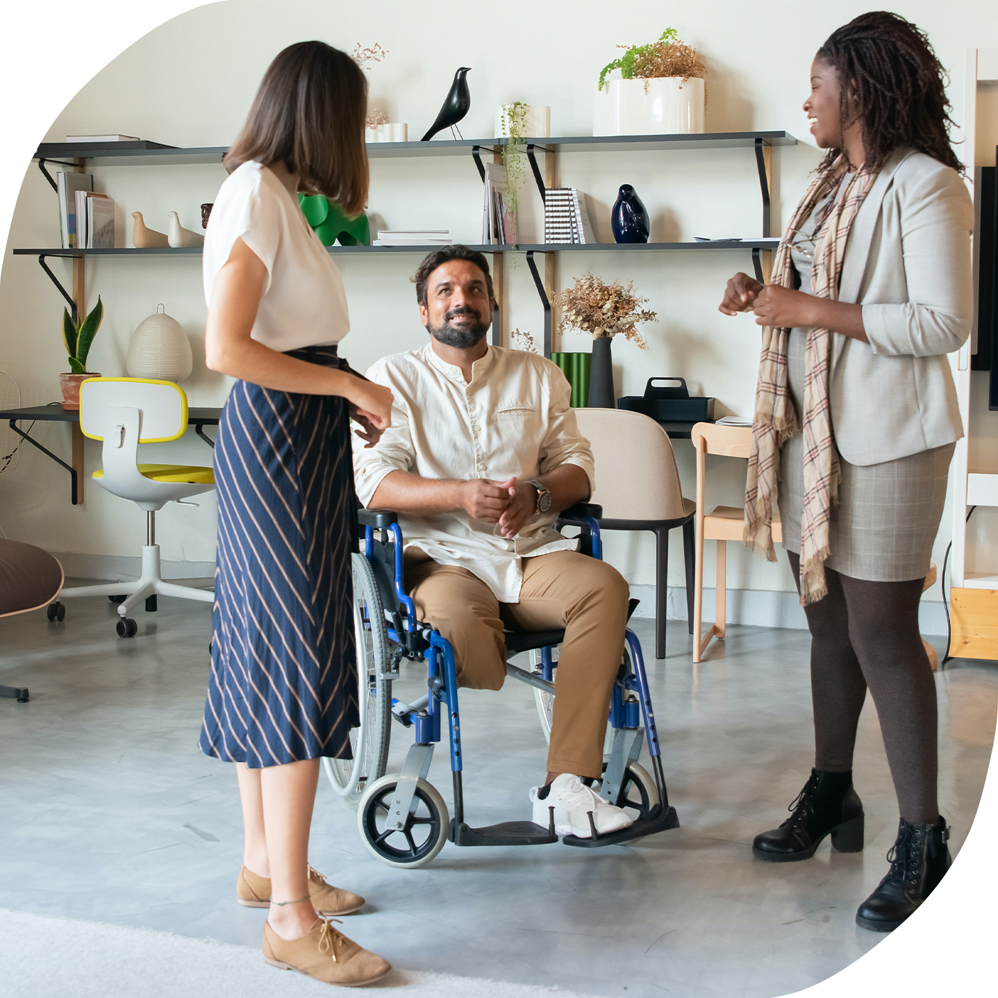 A photo of three co-workers, an Asian woman, a European man in a wheelchair, and a Black woman standing together and chatting at an office.