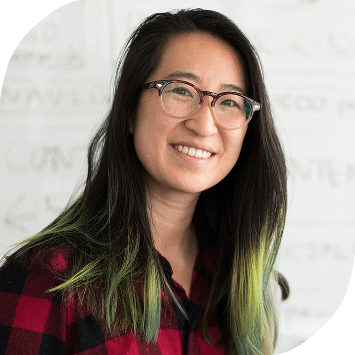 East Asian woman with long hair, glasses and a plaid shirt smiles in front of a whiteboard.