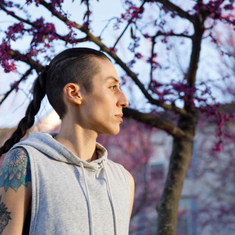 A transgender person wearing a sleeveless gray hoodie. They have dark hair that is shaved on the sides and worn in a ponytail. They are standing in the street, there are trees with pink blossoms behind them. They are looking off camera.