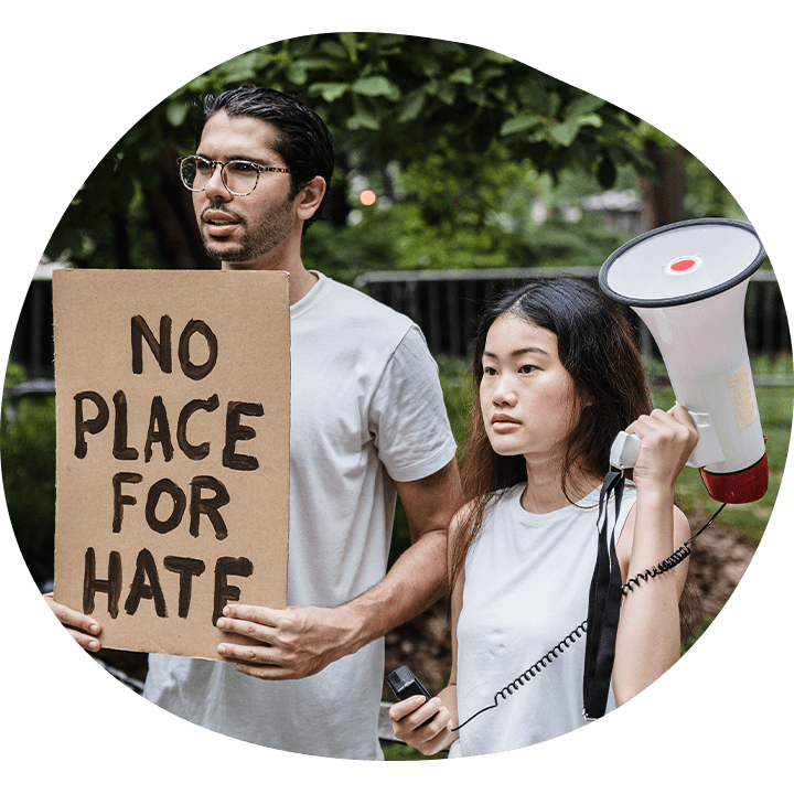 Man with medium skin tone, dark hair and glasses holds up a cardboard sign that says, "No place for hate." Beside him, an Asian woman with long black hair and a white tank top holds up a loudspeaker.