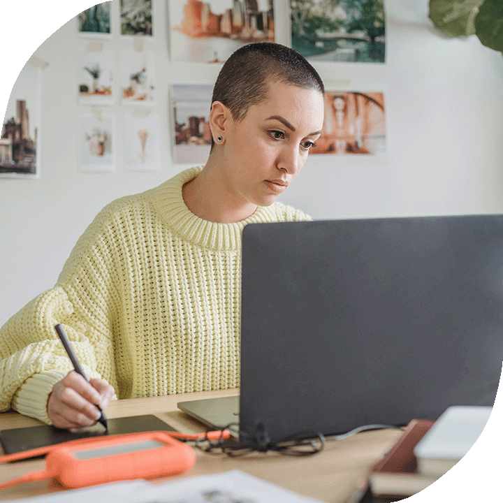 Woman with light skin tone and a shaved head is focused on a laptop screen as they use a tablet. They are wearing a light yellow jumper. The room is bright with natural light and posters and images are displayed on the back white wall.