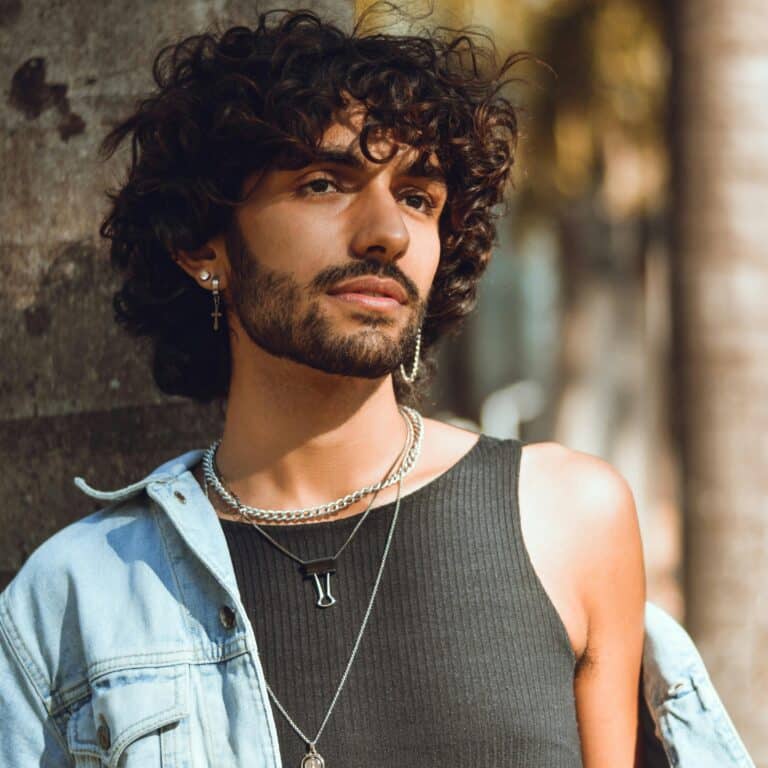 Young man with curly brown hair and beard wearing a tank top and jean jacket
