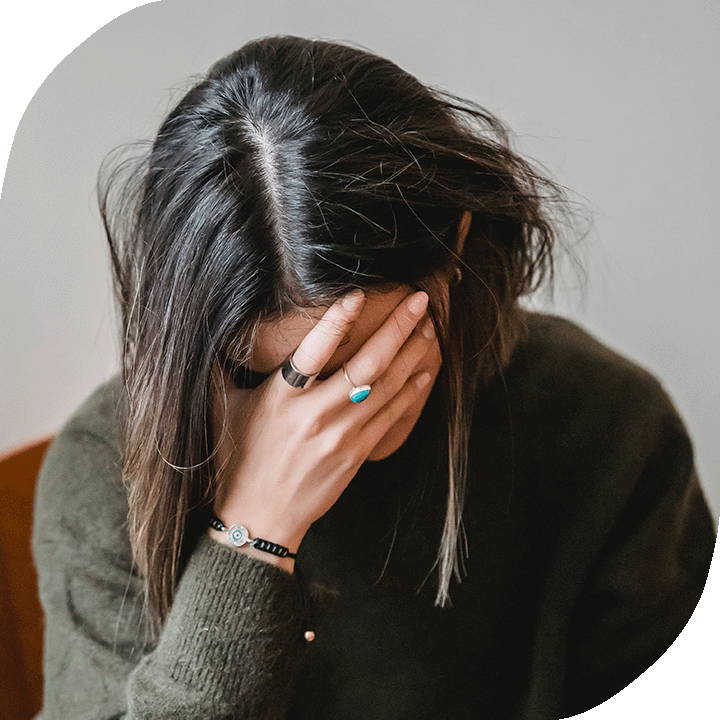 A distressed woman has her face in her hand. She has light skin tone, black hair and a dark green sweater.