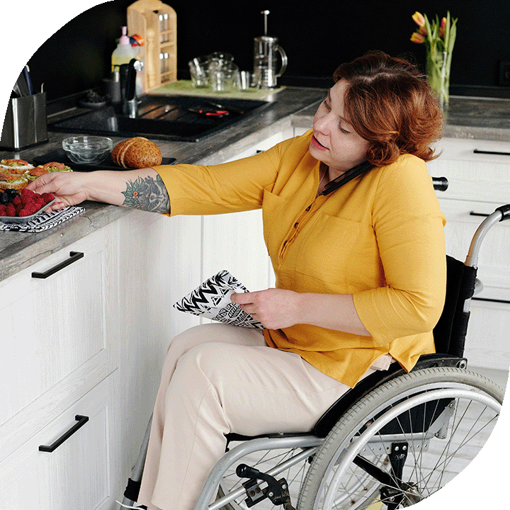 A woman goes about her kitchen in a wheelchair. She is white, has red hair, and wears a yellow top and beige pants.