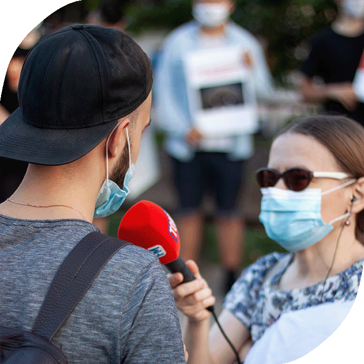 A white woman with sunglasses and a N95 mask interviews a man on the street. He is wearing a black cap, an N95 mask and a grey t-shirt.