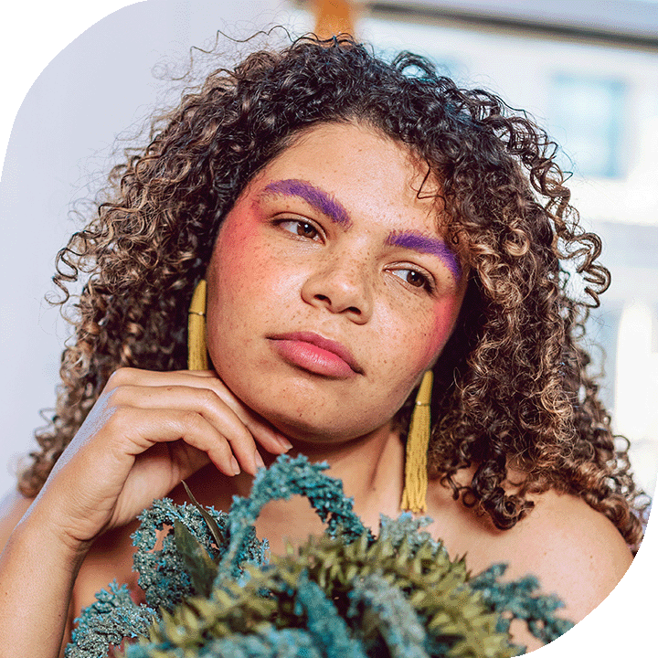 A woman with light-medium toned skin and brown curly air cocks her head to one side, contemplating. She wears bright colourful makeup and yellow tassel earrings.