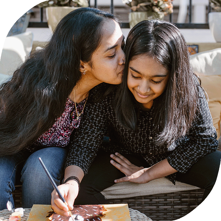 A mother kisses her daughter's cheek as her daughter smiles and draws a picture. Both have long black hair and medium-light skin tones.